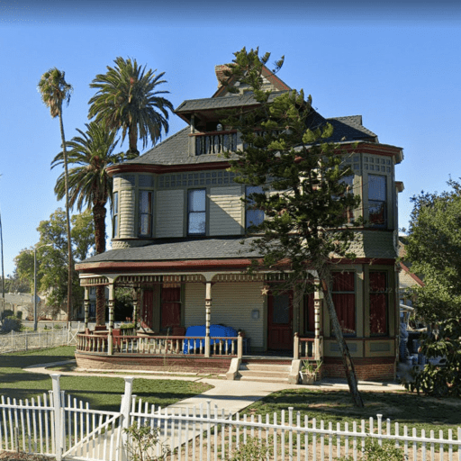 Historic Victorian Queen Anne-style house with wraparound porch, decorative wood trim, bay windows, white picket fence, and tall palm trees under a clear blue sky.